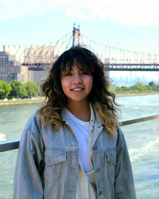 portrait of woman with dark hair standing in front of a bridge over water