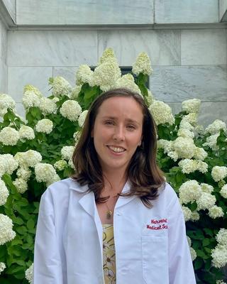 woman with brown hair in a a white coat standing in front of flowers