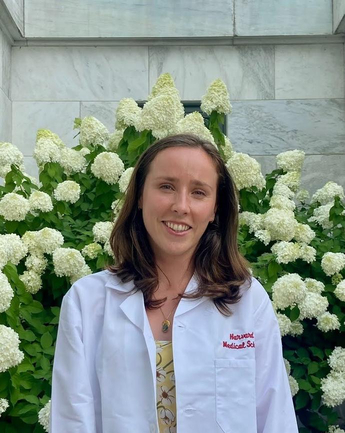 woman with brown hair in a a white coat standing in front of flowers