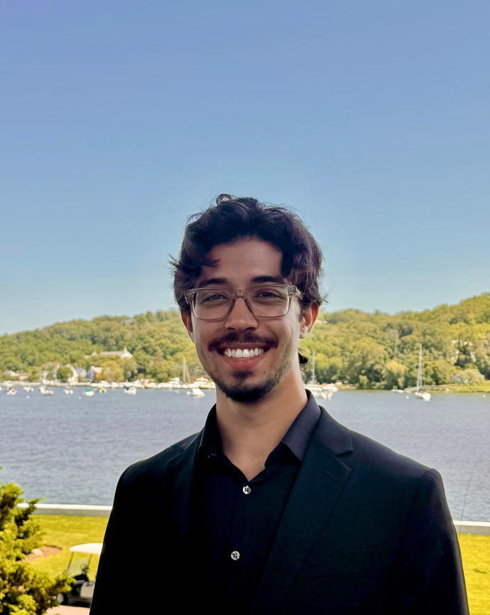 man with dark hair, glasses, and facial hair standing in front of a lake